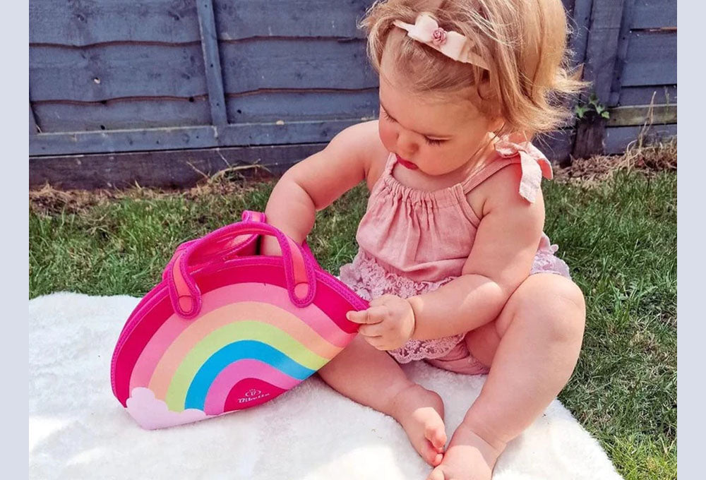 Little girl in a pink playsuit and bow in her hair, sitting on a blanket on the grass opening up a Bibetta rainbow lunch bag