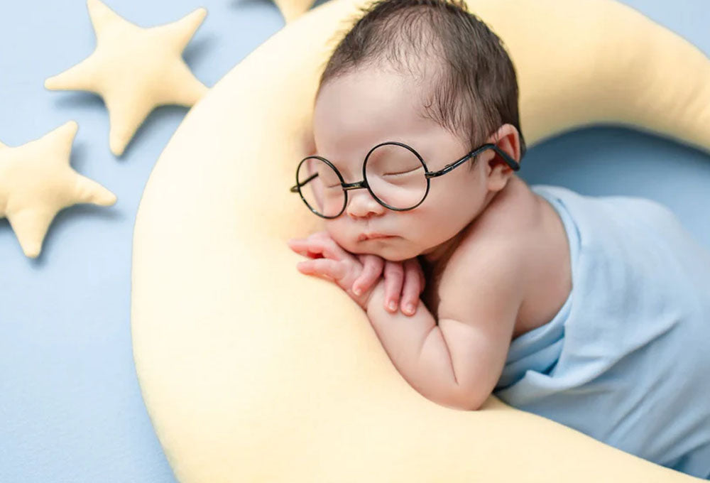 Baby sleeping or napping on a moon shaped pillow wrapped in a blue swaddle and wearing a pair of glasses.