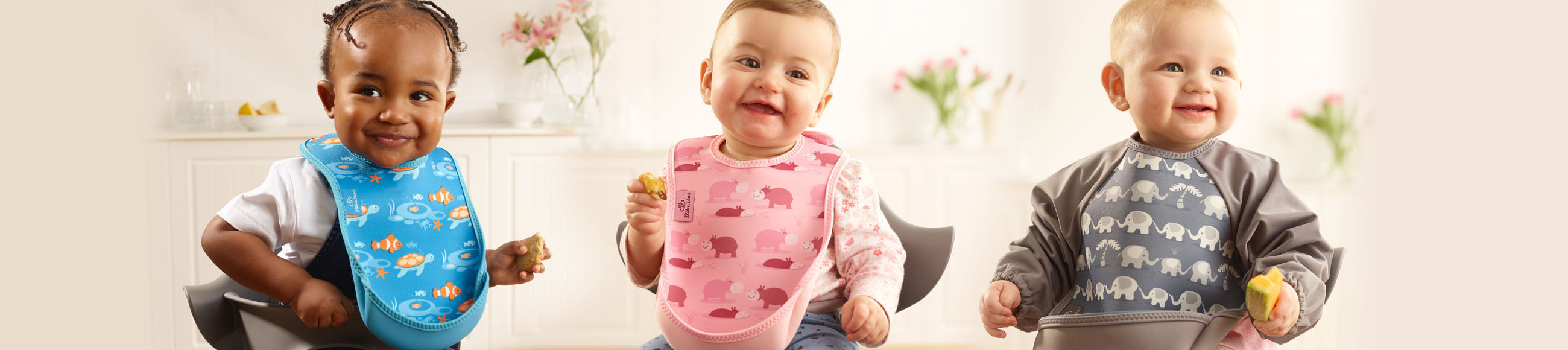 3 kids in Bibetta UltraBibs. 1st has a white t-shirt and blue bib with tropical fish pattern, 2nd has a long sleeved floral pink t-shirt on and pink bib with pink hippos pattern, 3rd has a long sleeved Ultrabib in grey and a light grey elephant pattern.