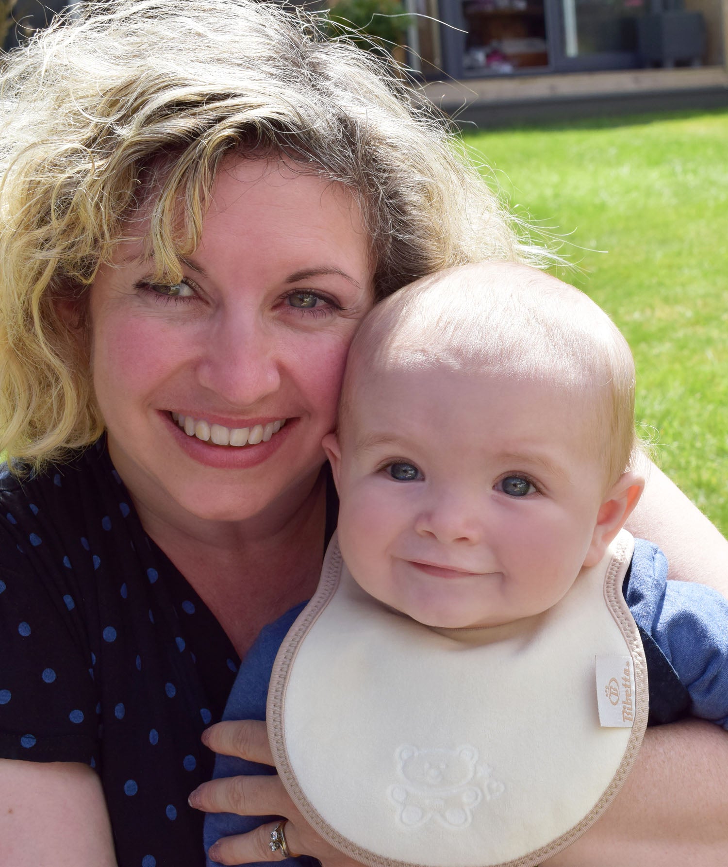 baby in a blue top wearing a bibetta cream and beige plush newborn bib with a teddy embossed into the front of the bib. The baby is being held by his mum.