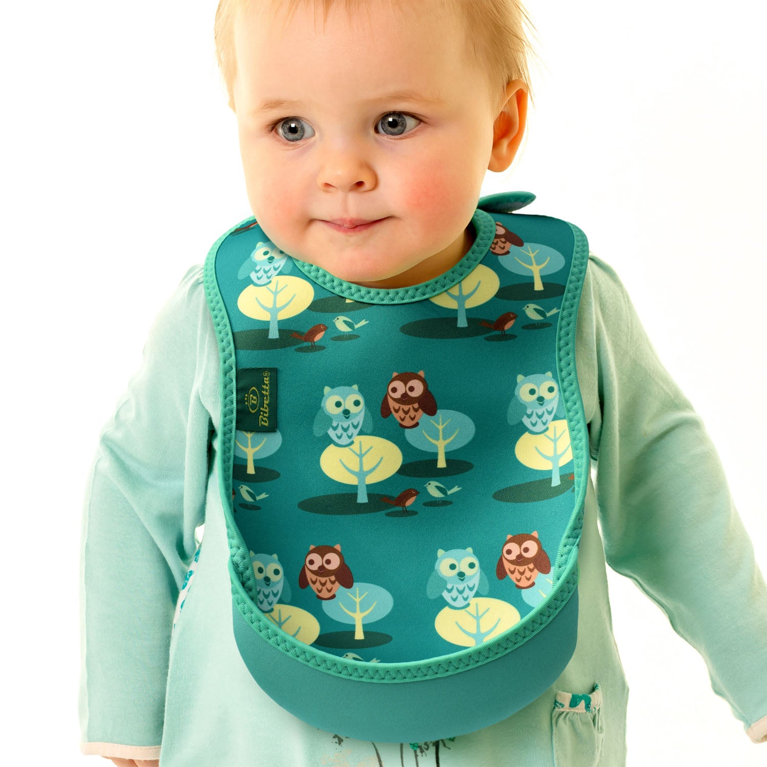 Baby wearing a Bibetta green weaning
bib with an owl and forest pattern, made of neoprene material, with the pocket flipped out to the front wearing a white and light green long sleeved dress