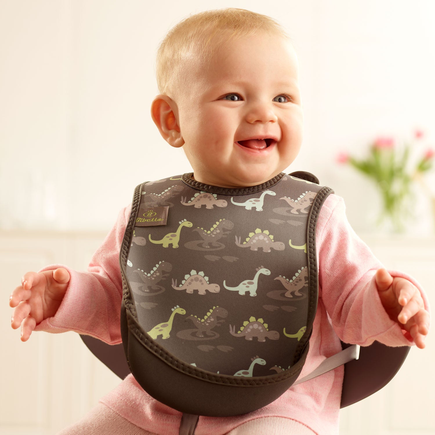 Baby wearing a Bibetta grey  weaning bib with a dinosaurs pattern, made of neoprene material, with the pocket flipped out to the front, sitting in a highchair wearing a pink long sleeved t-shirt and leggings