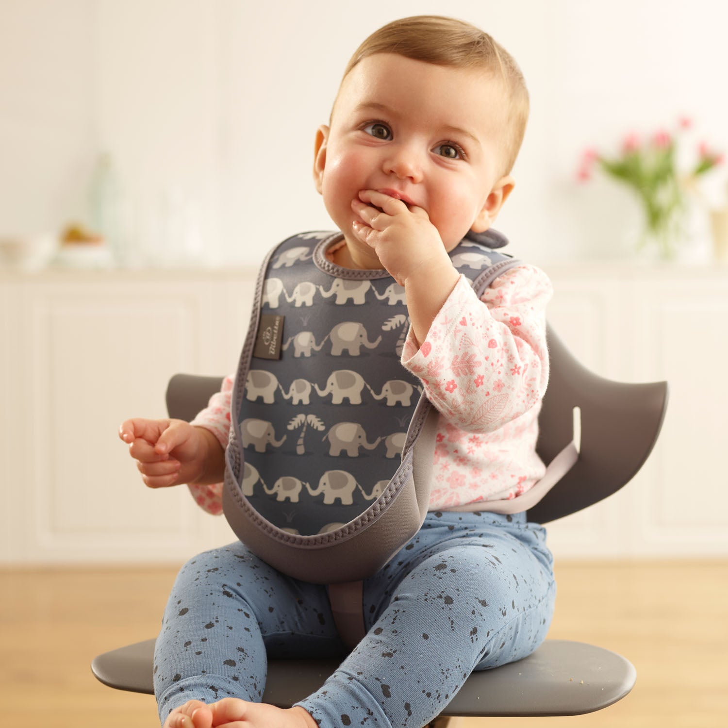 Baby wearing a Bibetta grey weaning 
 bib with an elephants pattern, made of neoprene material, with the pocket flipped out to the front, sitting in a highchair wearing a white and pink floral long sleeved t-shirt and blue patterned leggings
