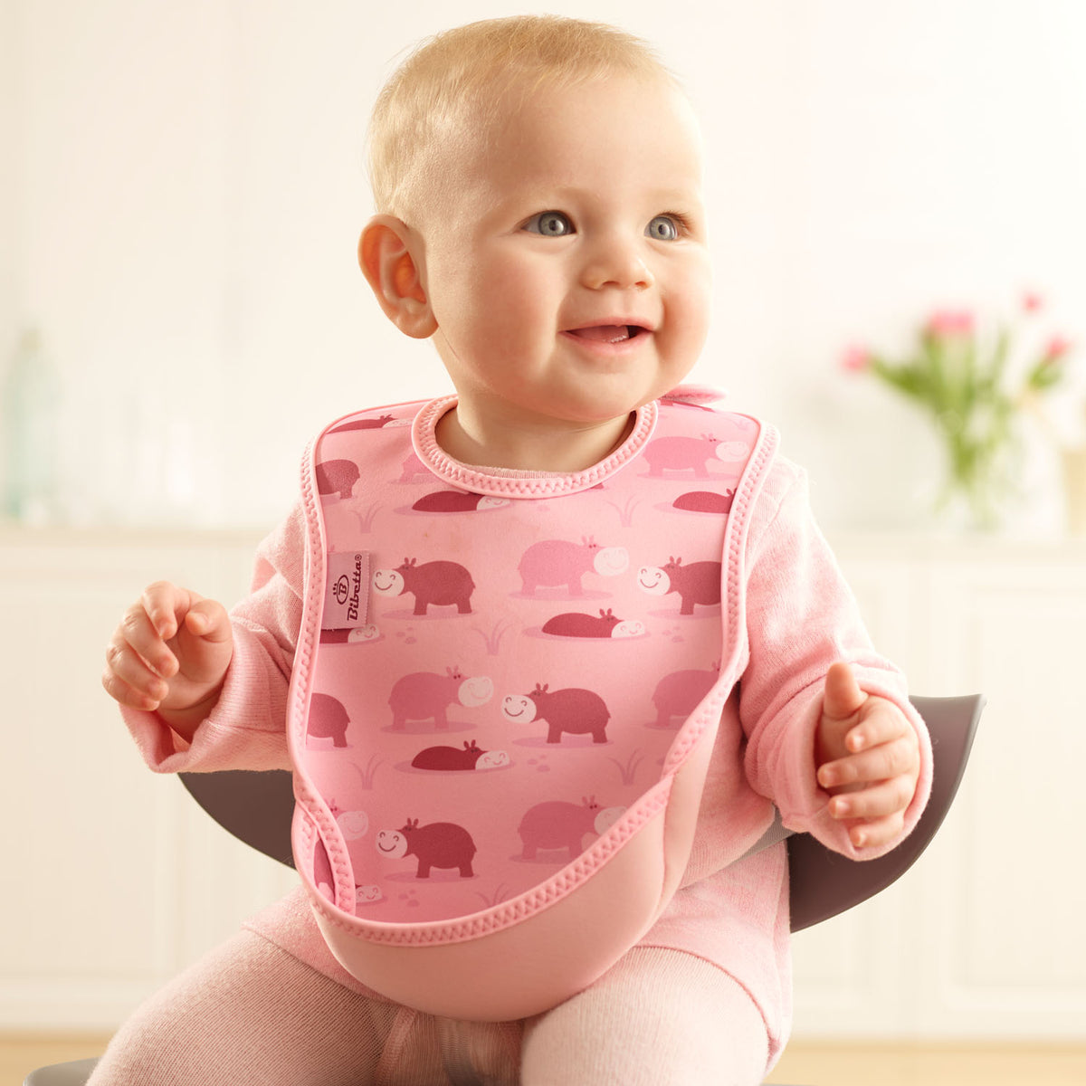 Baby wearing a Bibetta pink weaning bib with a hippos pattern, made of neoprene material, with the pocket flipped out to the front, sitting in a highchair wearing a pink long sleeved t-shirt and leggings