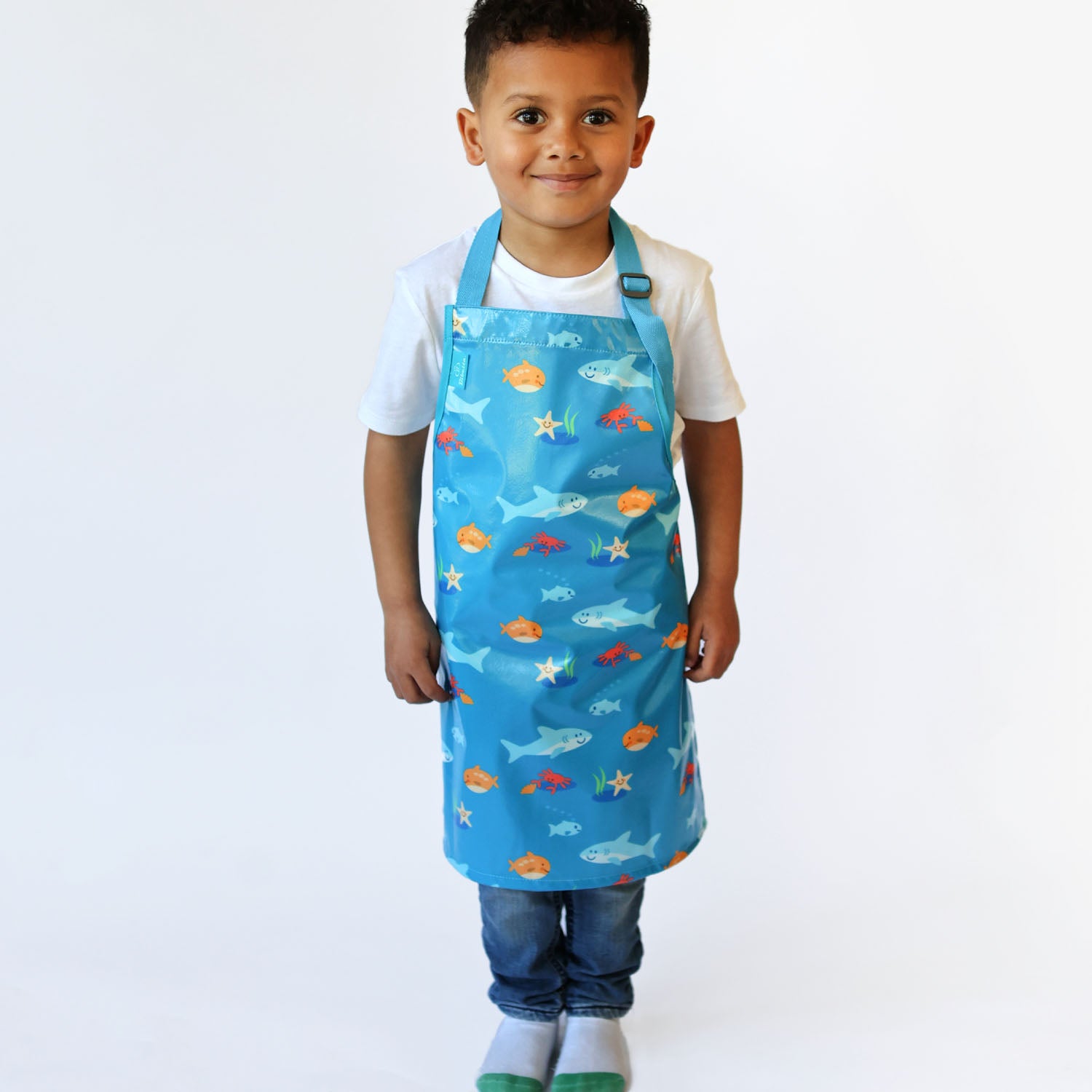 a smiling boy standing wearing a Bibetta Wipeezee childs apron in turquoise sea creatures pattern. He is wearing a white t-shirt and blue jeans with white and green socks.
