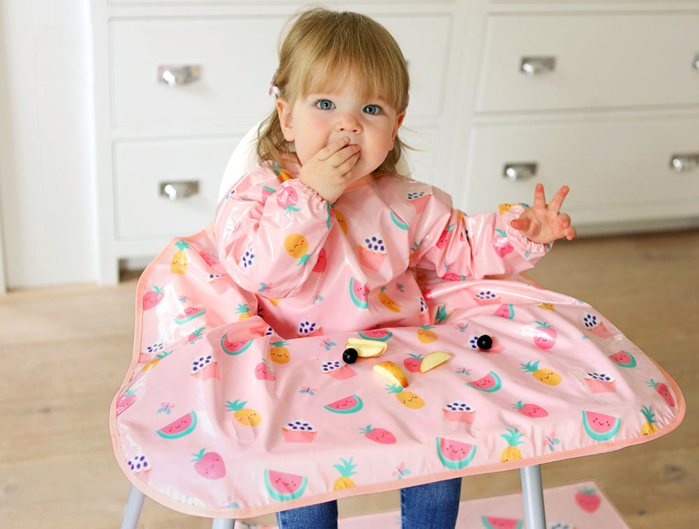 A happy toddler child sat in a highchair wearing a Bibetta Wipeezee pink Yummy Treats print coverall baby bib that covers the highchair table.