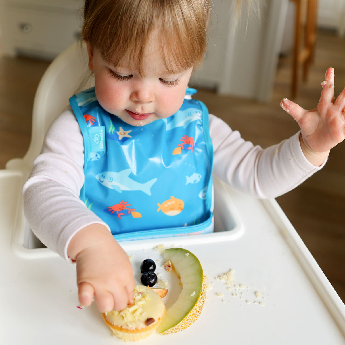 a toddler seated in a white ikea highchair wearing a Bibetta wipeezee weaning bib in turquoise sea creatures with the pocket folded to the front. The baby is dressed in a white long sleeved and is playing with the food on the highchair tray.