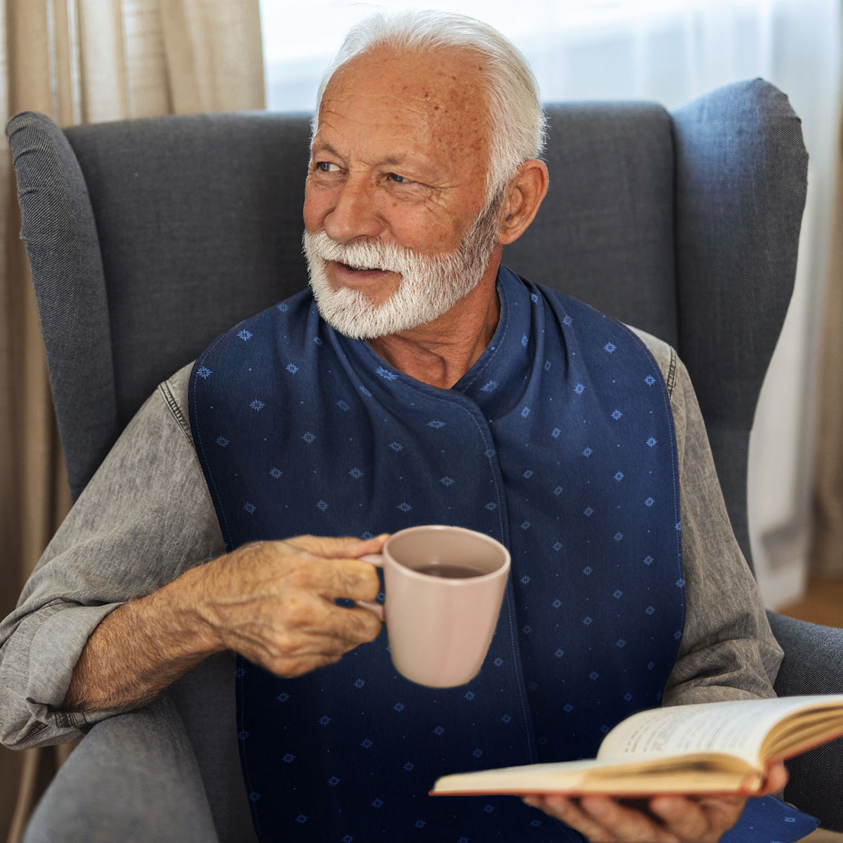 Older gentleman sitting in a chair holding a cup and book, wearing a navy blue patterned straight scarf clothing protector