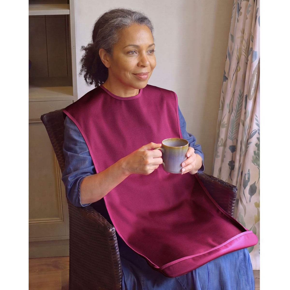 A woman sitting in a dark armchair indoors, wearing a Care Designs large tabard style bib in burgundy over her blue dress and holding a small ceramic mug.