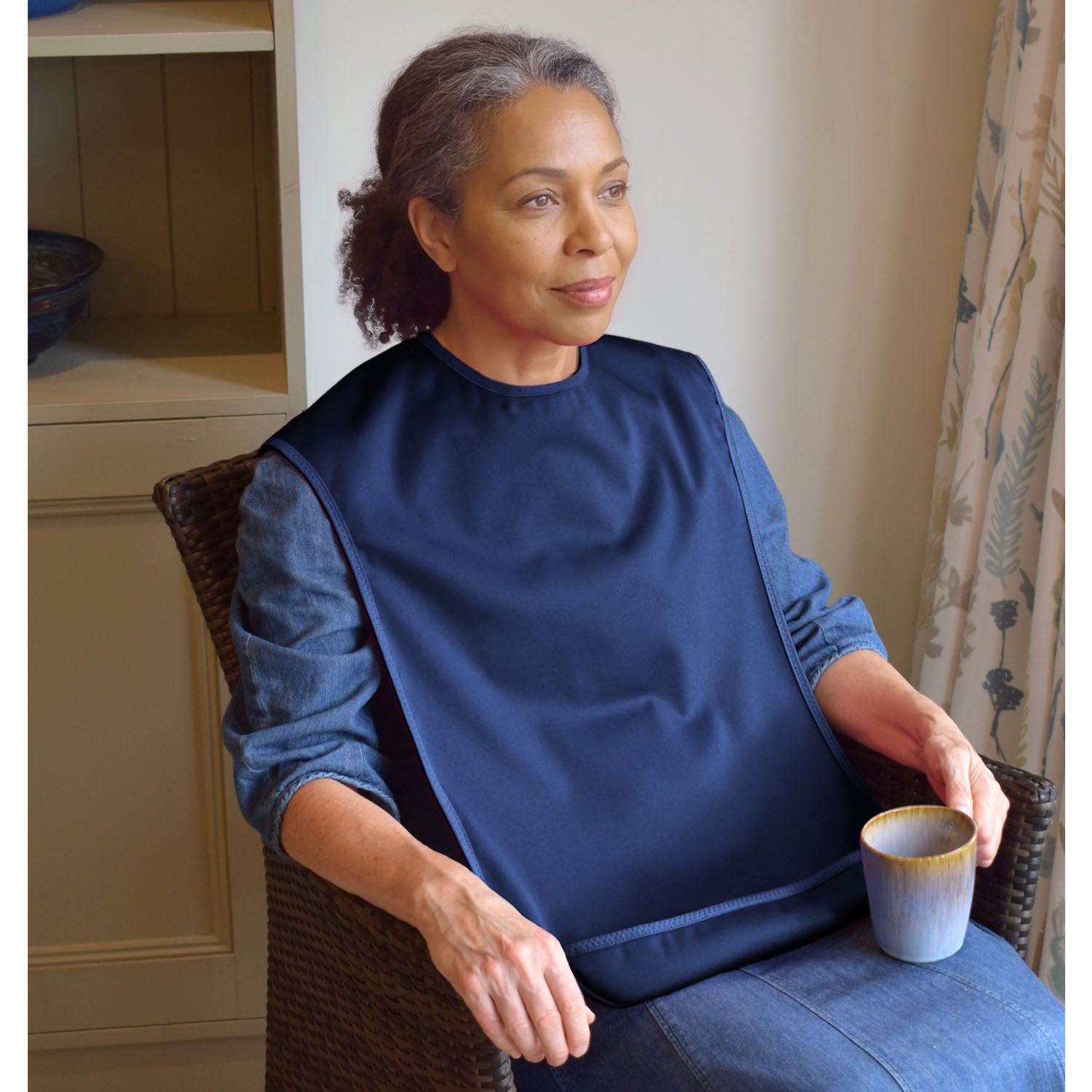 A woman sitting in a wicker armchair wearing a Care Designs navy blue tabard style bib over a denim shirt, holding a small cup in a home interior setting.
