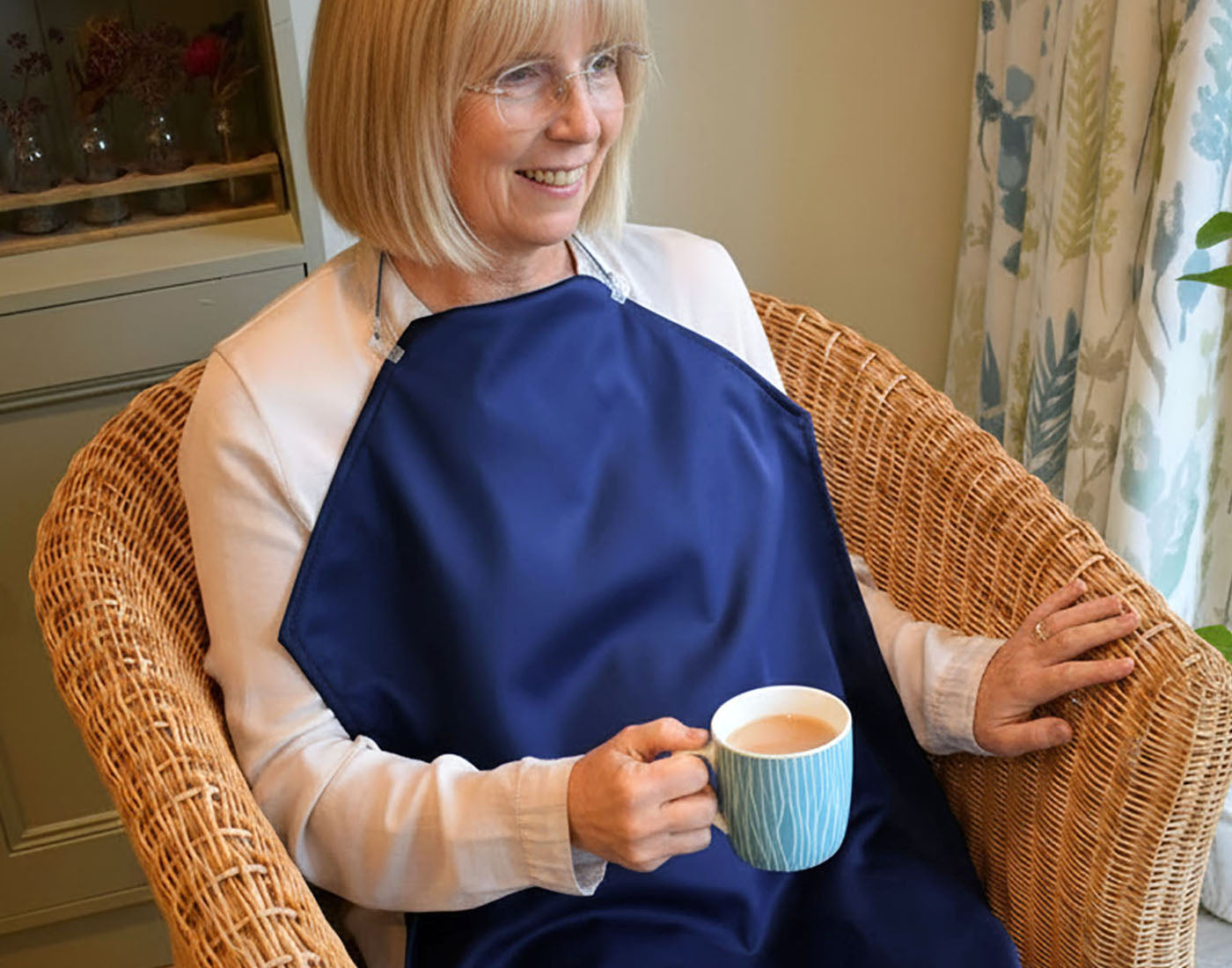 Senior woman sitting in a chair wearing a navy blue, long napkin, apron-style, adult bib clothing protector whilst holding a mug of tea.