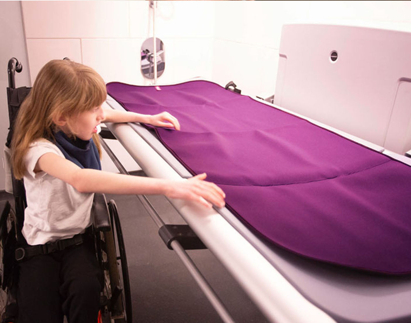 A young girl sitting in a wheelchair next to a changing spaces bed in an accessible toilet with a Care Designs aubergine, adult changing mat on top of the bed or changing table.