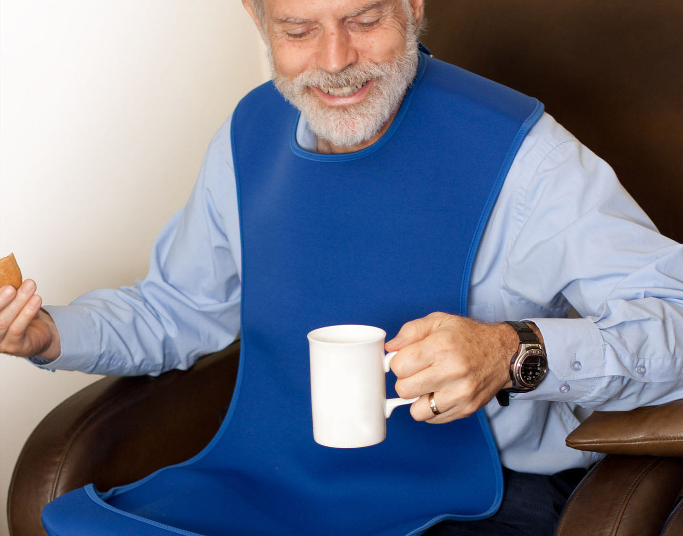 Elderly man, senior gentleman sitting in a chair holding a cup of tea and wearing a light blue shirt protectred by a Care Designs XL blue Tabard adult bib clothing protector with a crumb catcher pocket.
