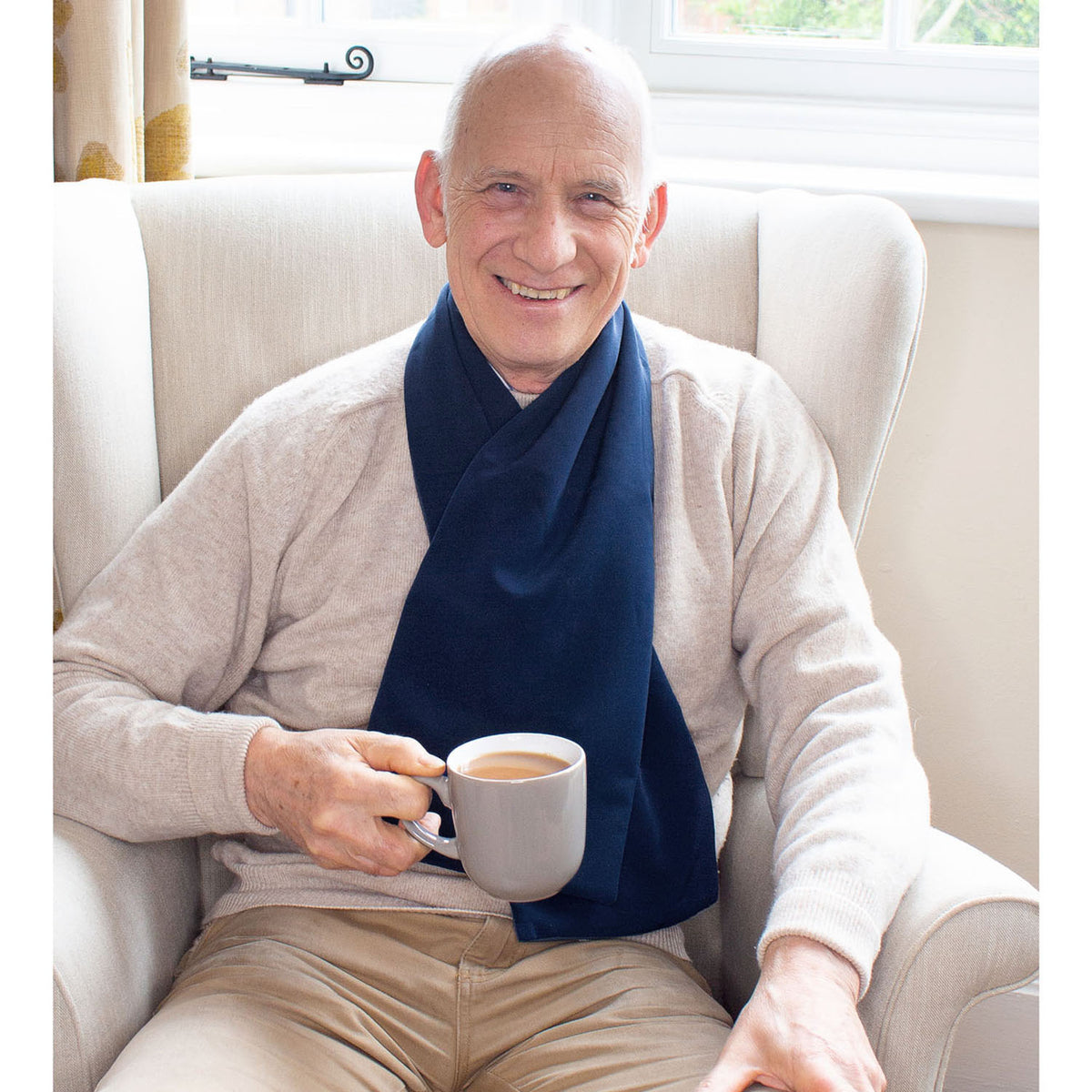 Man sitting in a chair holding a cup of coffee, wearing a beige sweater and navy cross scarf clothing protector.