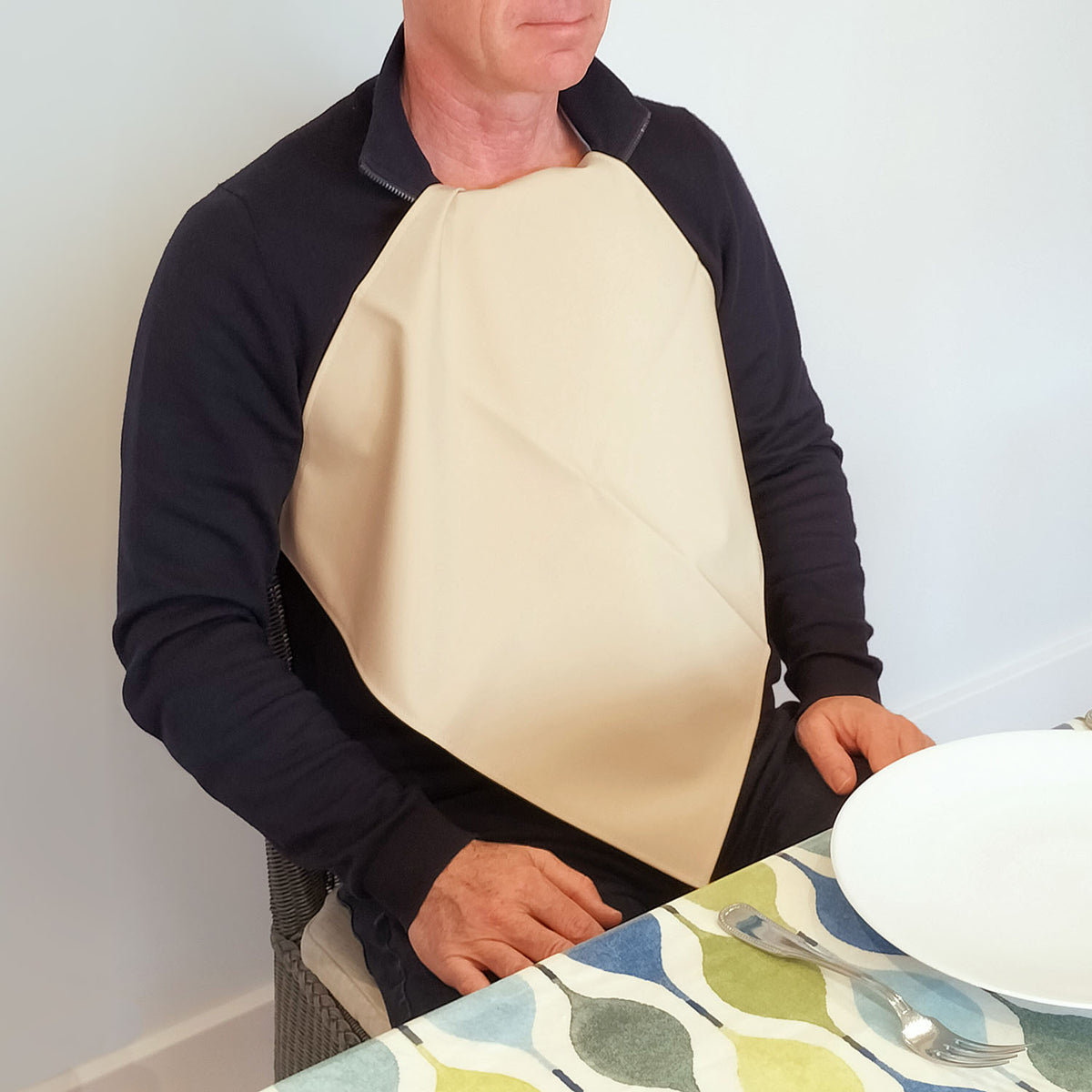 Man wearing a Care Designs cream waterproof napkin over a black shirt at a dining table with a colorful placemat.