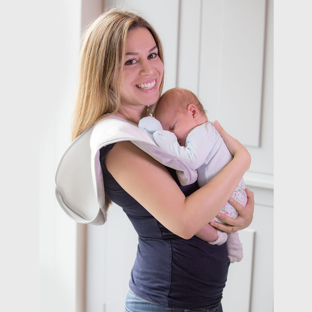 ady with brown hair wearing a long blue t-shirt holding a baby against her shoulder in a light blue top and flower leggings and pick socks with a Bibetta white and grey burp cloth