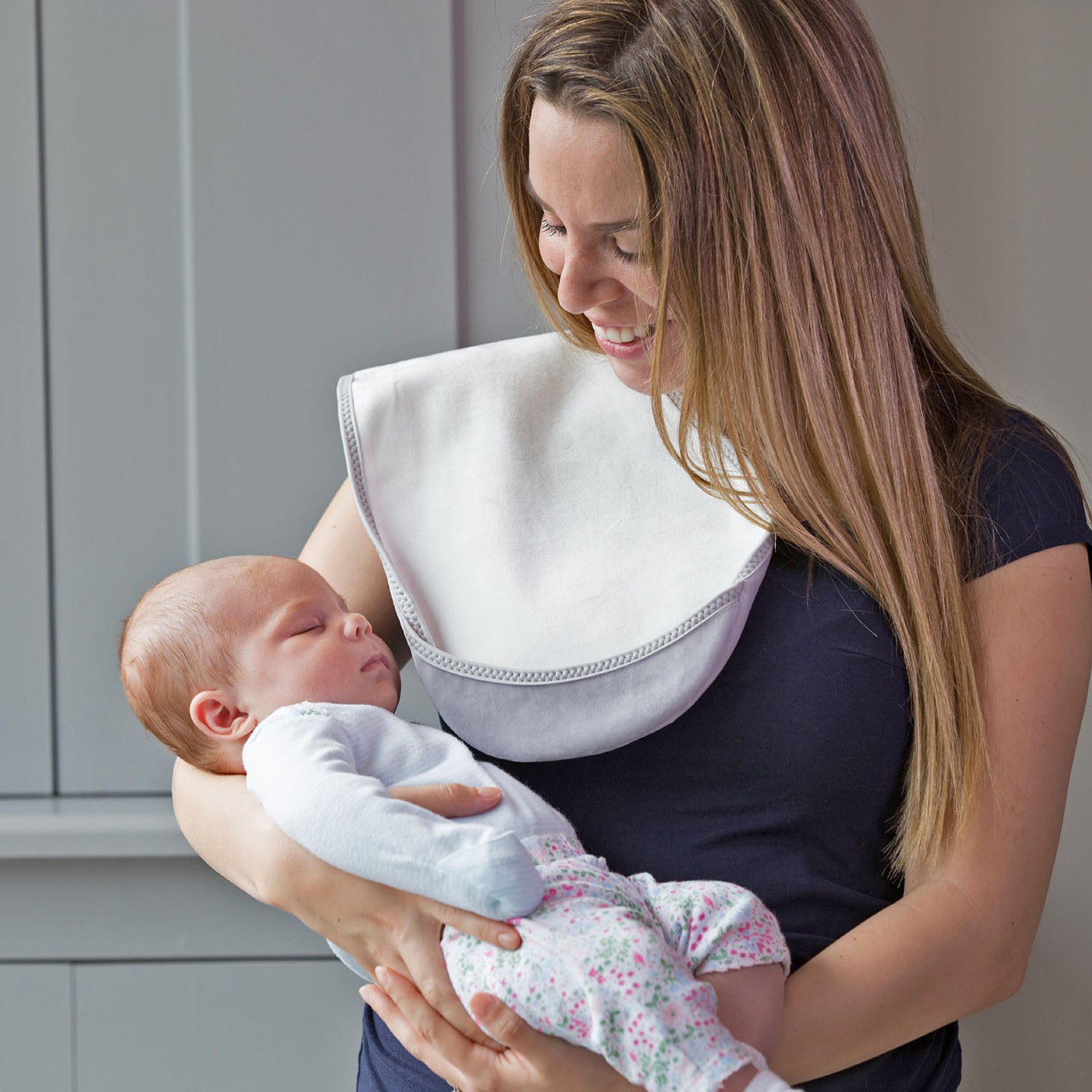 lady with brown hair wearing a long blue t-shirt holding a baby in a light blue top and flower leggings and pick socks in her arms  with a Bibetta white and grey burp cloth on her shoulder