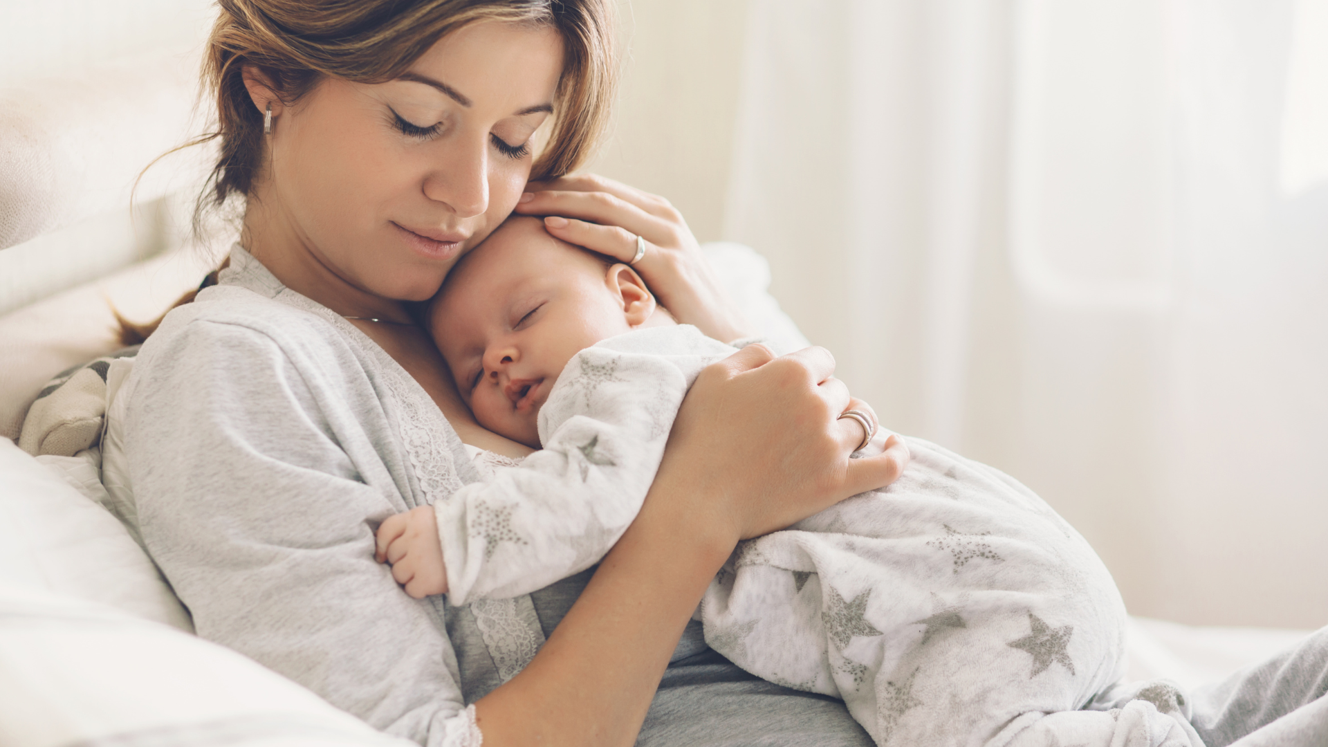 A lady with brown hair lying back on a bed wearing a white top snuggling a sleeping baby on her chest. The baby is wearing a white sleepsuit with silver stars on it.