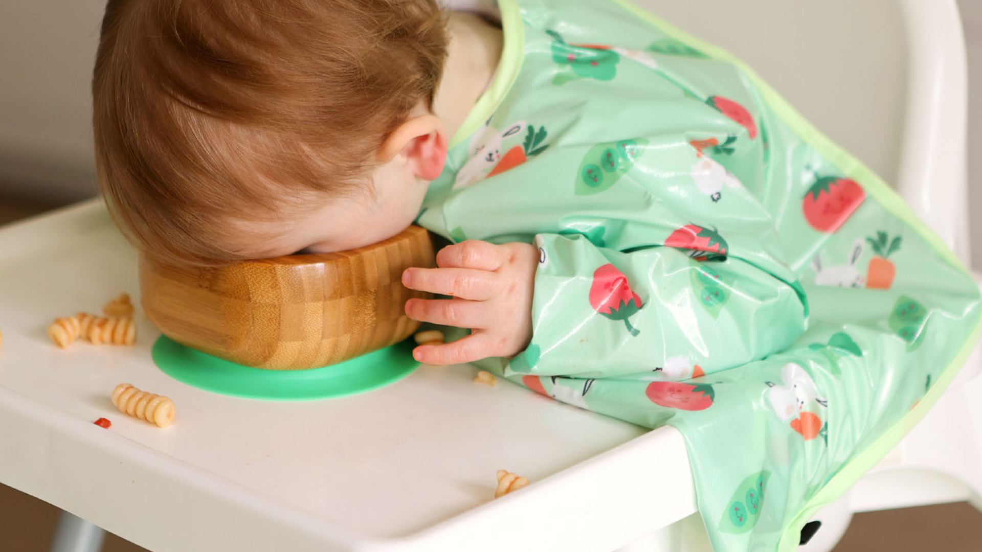 Little boy wearing a green bunny and veg wipeezee coverall sitting in a highchair with this face in a bowl on the highchair tray.
