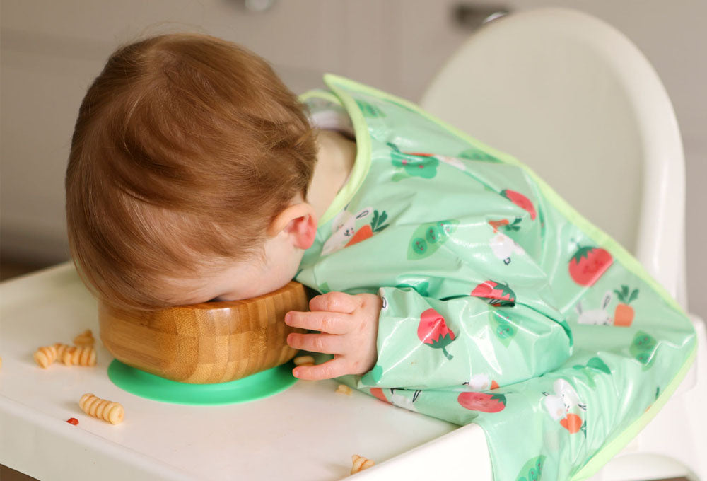 Little boy wearing a green bunny and veg wipeezee coverall sitting in a highchair with this face in a bowl on the highchair tray.