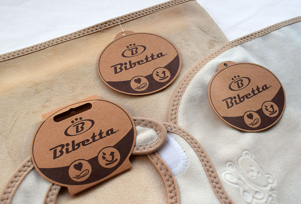 Bibetta's newborn plush products in cream and beige (top of baby bib, corner of baby mat and section of the back of the burp cloth) highlighting the card hangers (plain card with sepa colour print on them) for each products