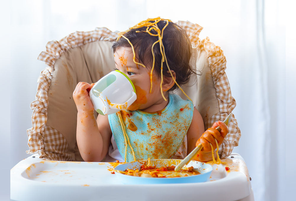 Baby sat in a highchair covered in spaghetti and tomato sauce. The baby is wearing a bib and drinking out of a cup whilst eating food on a plate using a spoon.