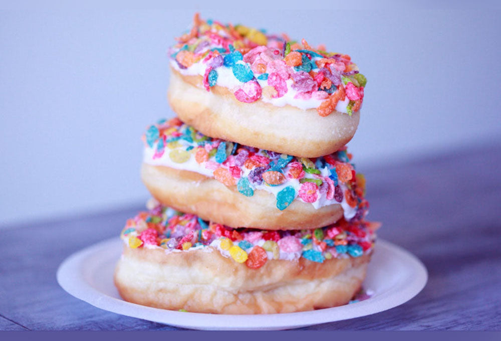 Three doughnuts with icing and different colours of sugar sprinkles on them stacked on top of each other on a white plate