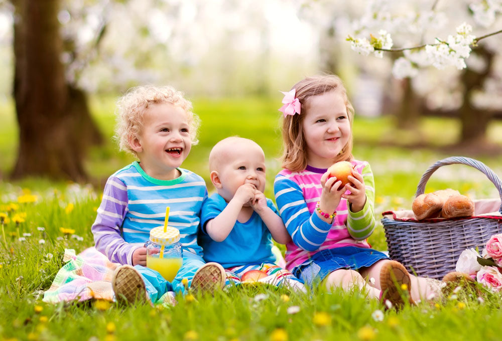 Three children, a toddler, baby and older child sitting on a picnic mat on the grass eating and drinking. There is a picnic basket next to them with food in it.