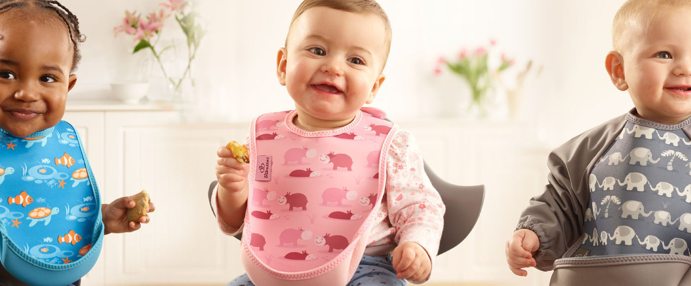 3 kids in Bibetta UltraBibs. 1st has a white t-shirt and blue bib with tropical fish pattern, 2nd has a long sleeved floral pink t-shirt on and pink bib with pink hippos pattern, 3rd has a long sleeved Ultrabib in grey and a light grey elephant pattern.
