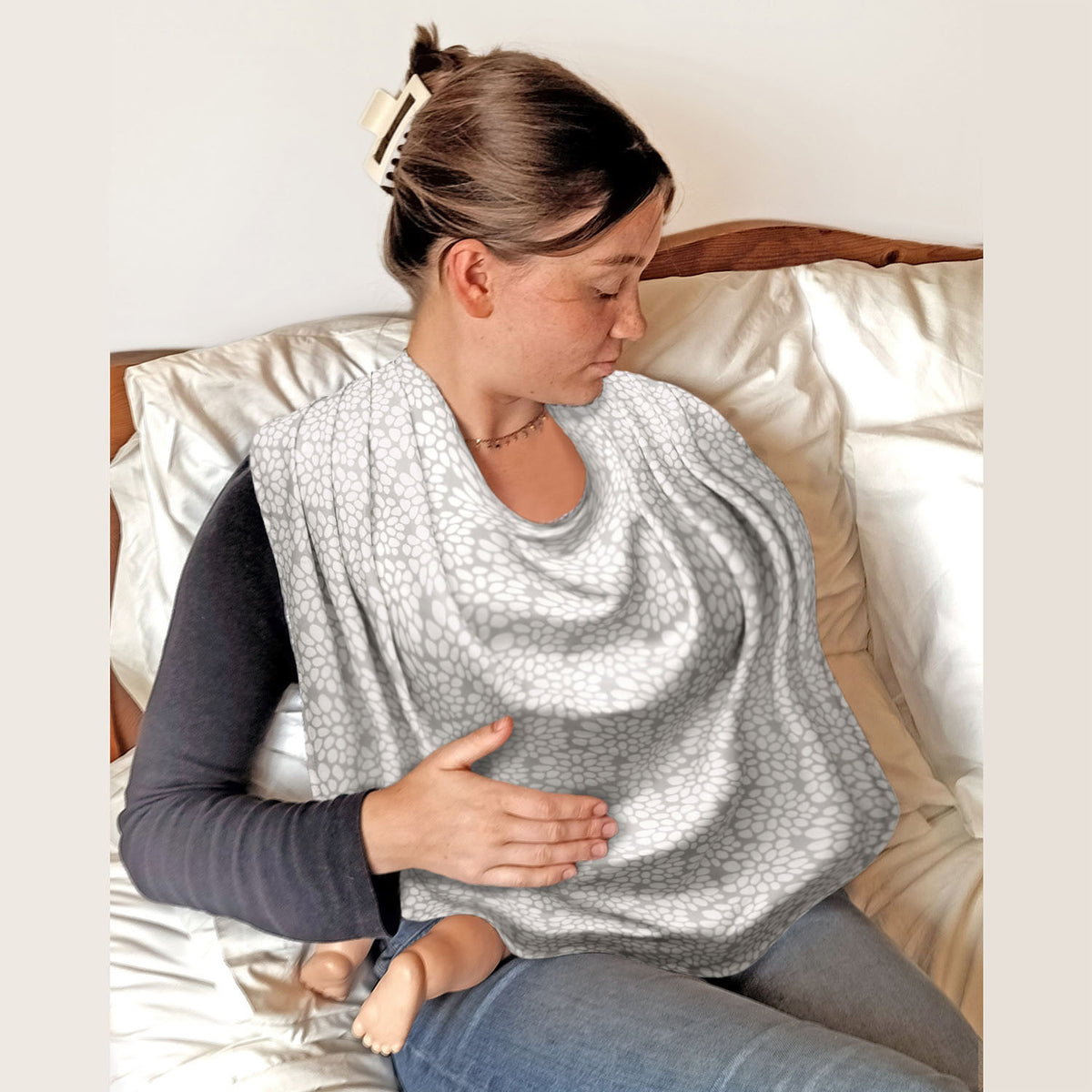 Woman sitting on a bed wearing a Bibetta Breastfeeding cover over her shoulders with her baby underneath