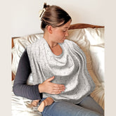 Woman sitting on a bed wearing a Bibetta Breastfeeding cover over her shoulders with her baby underneath
