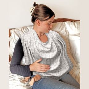 Woman sitting on a bed wearing a Bibetta Breastfeeding cover over her shoulders with her baby underneath