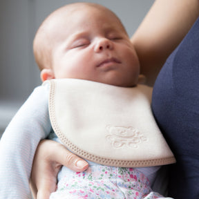 baby in a light blue top and flowery leggings wearing a bibetta cream and beige plush newborn bib with a teddy embossed into the front of the bib. The baby is being held by her mum.