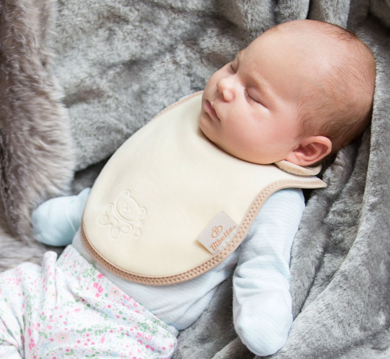 baby in a light blue top and flowery leggings wearing a bibetta cream and beige plush newborn bib with a teddy embossed into the front of the bib. The baby is lying on a grey faux fur blanket