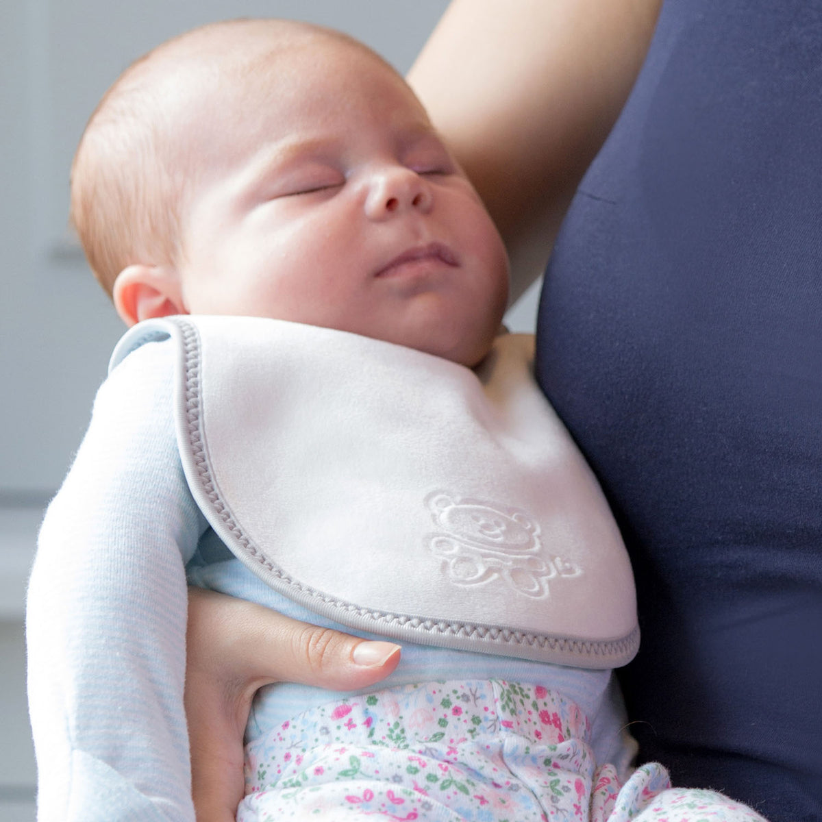 baby in a light blue top and flowery leggings wearing a bibetta white and grey plush newborn bib with a teddy embossed into the front of the bib. The baby is being held by her mum.