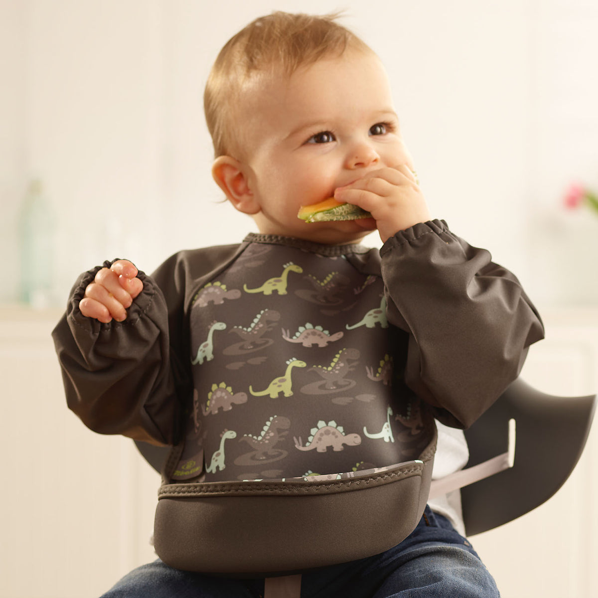 Baby boy wearing a Bibetta grey  weaning bib with sleeves with a dinosaurs pattern, made of neoprene material, with the pocket flipped out to the front, sitting in a highchair eating a melon wearing denim trousers