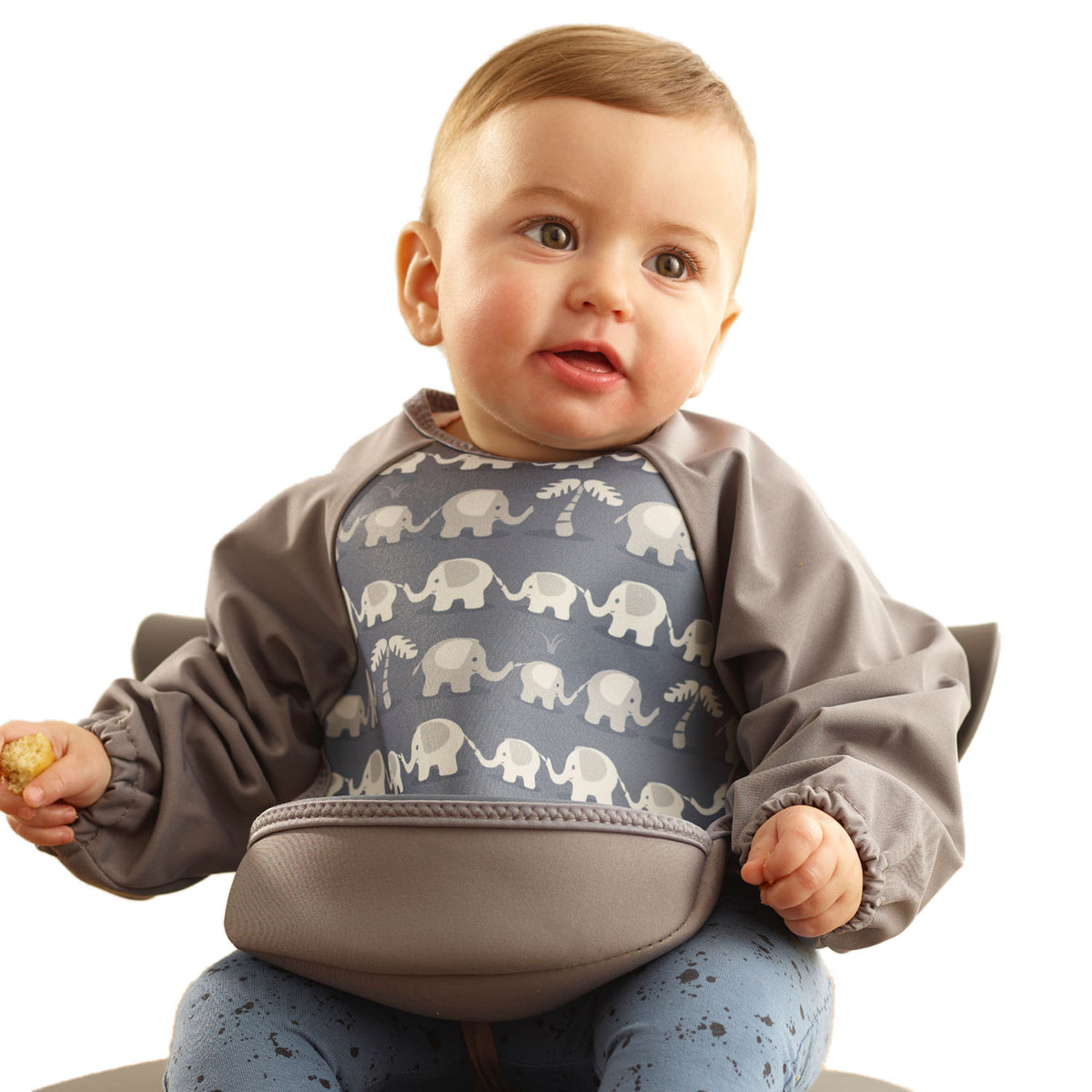 Baby girl wearing a Bibetta grey  weaning bib with sleeves with a dinosaurs pattern, made of neoprene material, with the pocket flipped out to the front, sitting in a highchair eating a biscuit wearing blue spotty leggings