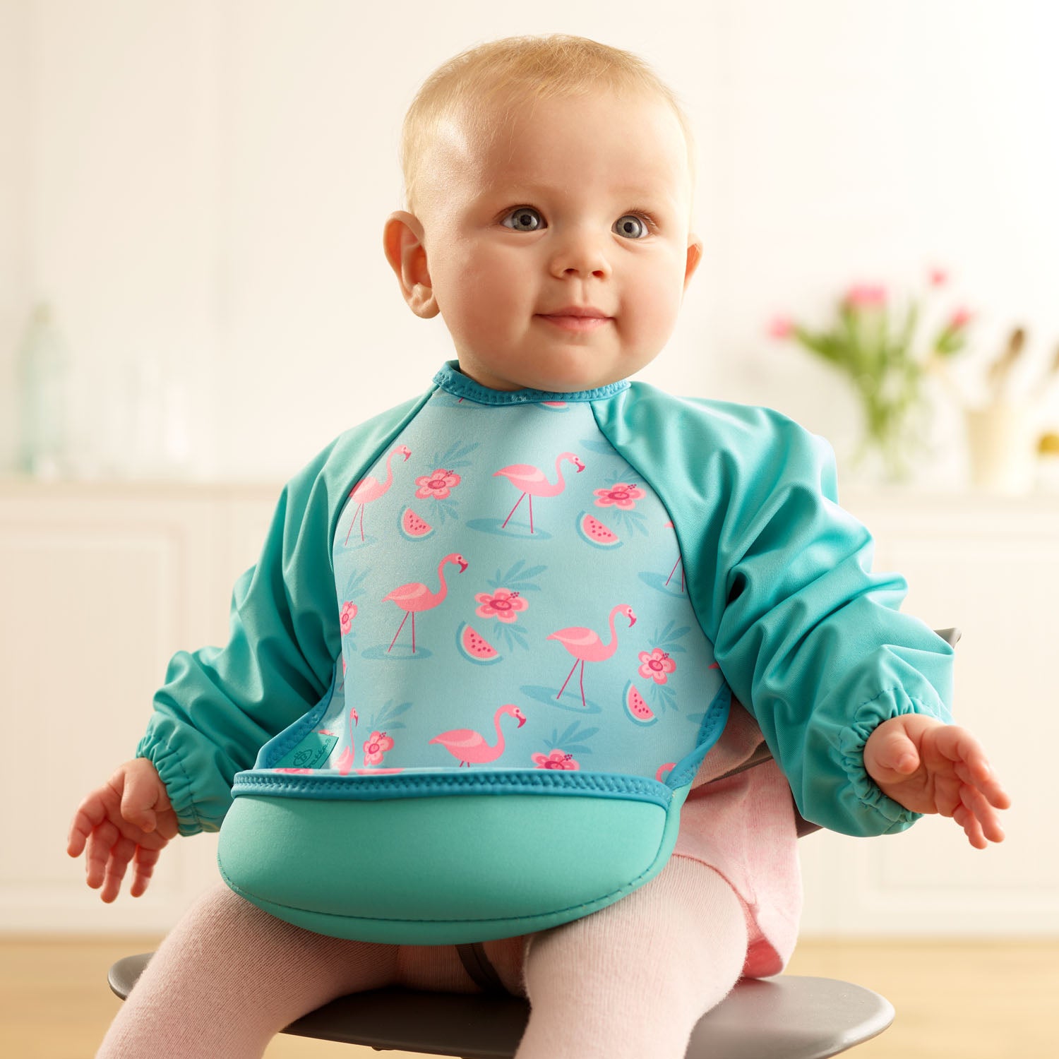 Baby girl wearing a Bibetta turquoise green weaning bib with sleeves with a pink flamingos pattern, made of neoprene material, with the pocket flipped out to the front, sitting in a highchair wearing a pink dress and tights