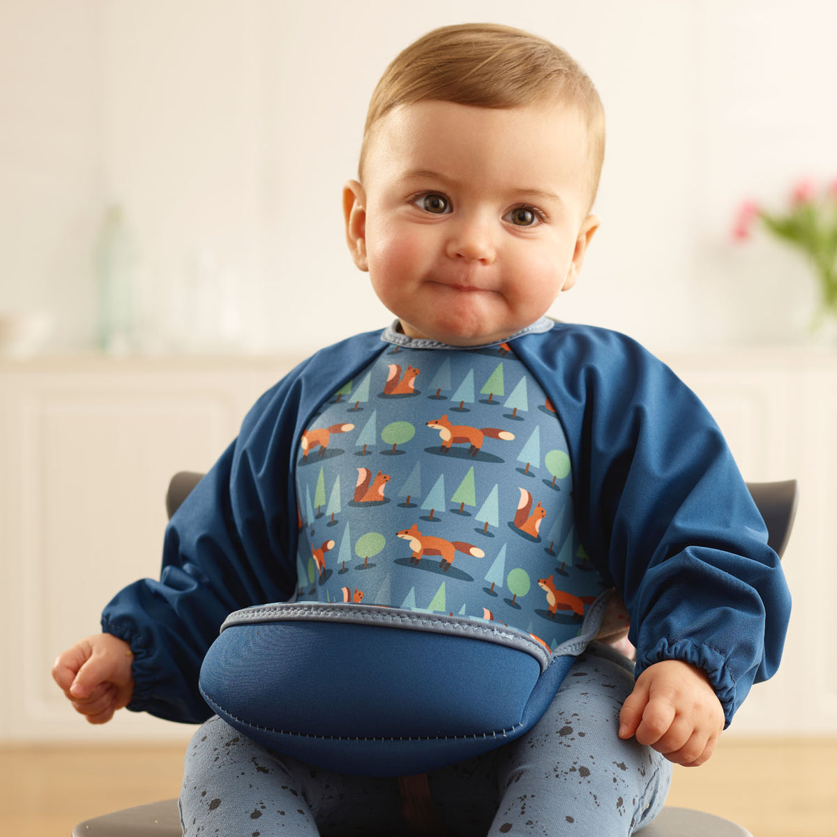 Baby girl wearing a Bibetta blue weaning bib with sleeves with a fox, squirrels and trees pattern, made of neoprene material, with the pocket flipped out to the front, sitting in a highchair wearing a blue spotty leggings