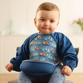 Baby girl wearing a Bibetta blue weaning bib with sleeves with a fox, squirrels and trees pattern, made of neoprene material, with the pocket flipped out to the front, sitting in a highchair wearing a blue spotty leggings