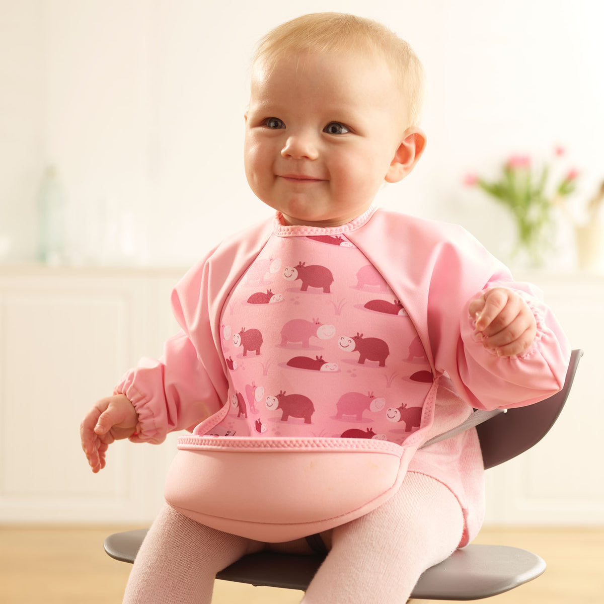 Baby girl wearing a Bibetta pink weaning bib with sleeves with a hippos pattern, made of neoprene material, with the pocket flipped out to the front, sitting in a highchair wearing a pink dress and tights.