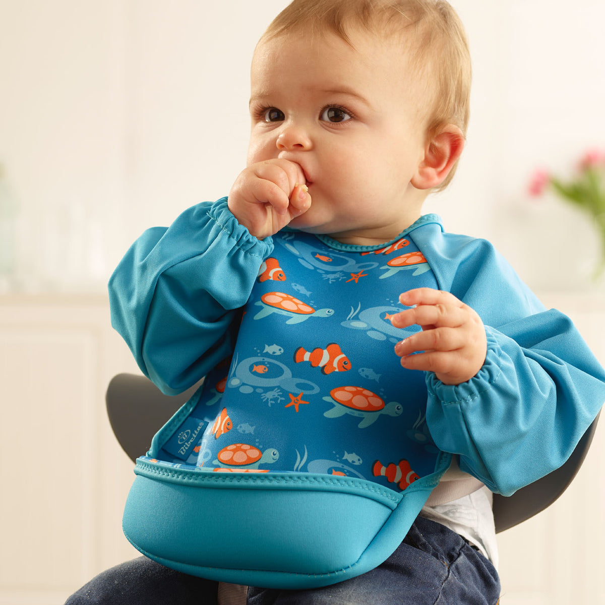 Baby boy wearing a Bibetta turquoise blue weaning bib with sleeves with orange fish and turtle pattern, made of neoprene material, with the pocket flipped out to the front, sitting in a highchair wearing a white t-shirt and denim jeans.