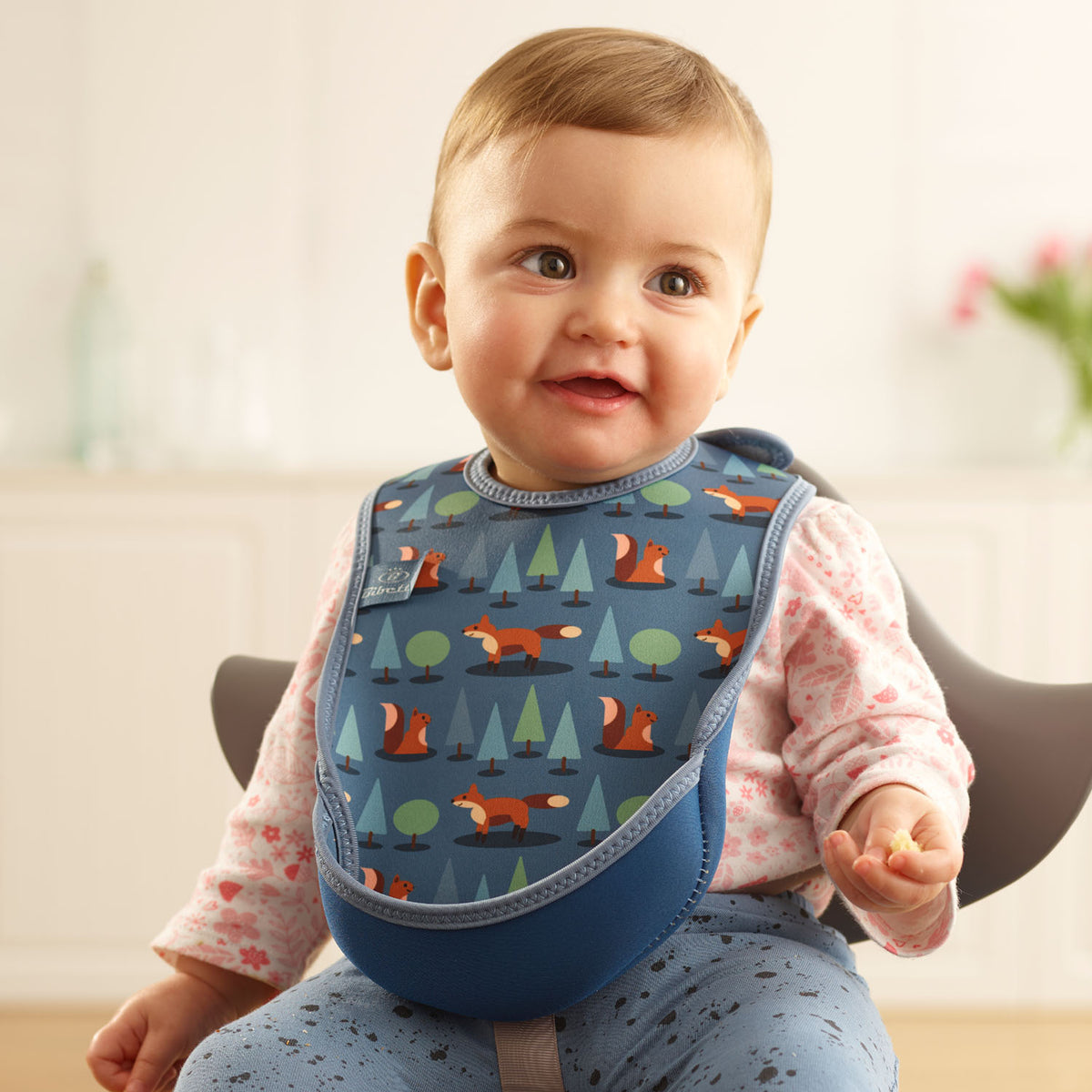 Baby wearing a Bibetta blue weaning 
 bib with a fox and forest pattern, made of neoprene material, with the pocket flipped out to the front, sitting in a highchair wearing a white and pink floral long sleeved t-shirt and blue patterned leggings 