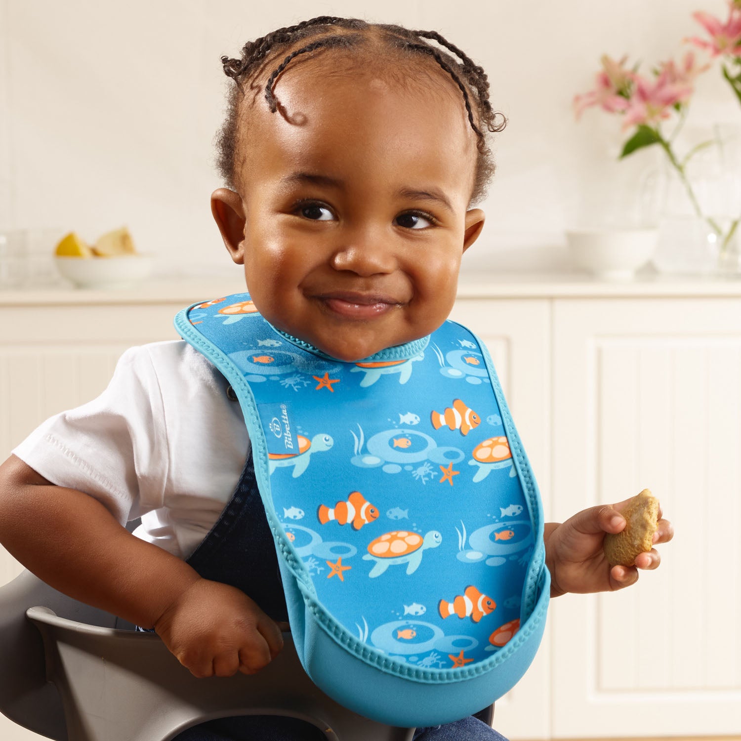 Baby wearing a Bibetta blue weaning bib with a tropical fish pattern, made of neoprene material, with the pocket flipped out to the front, sitting in a highchair wearing a white short sleeved t-shirt and denim dungarees