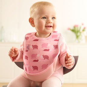 Baby wearing a Bibetta pink weaning bib with a hippos pattern, made of neoprene material, with the pocket flipped out to the front, sitting in a highchair wearing a pink long sleeved t-shirt and leggings 