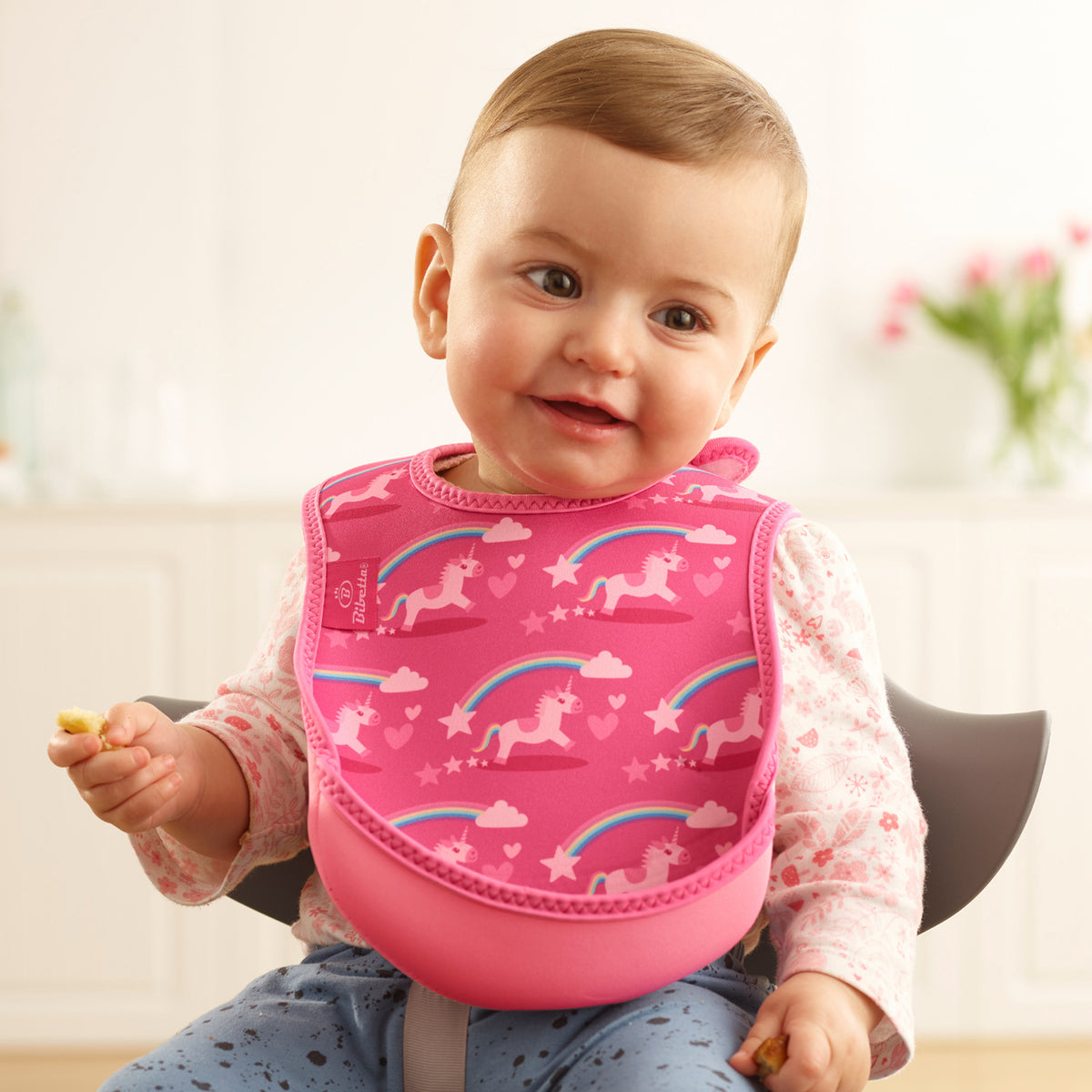 Baby wearing a Bibetta pink weaning bib with an unicorns pattern, made of neoprene material, with the pocket flipped out to the front, sitting in a highchair wearing a white and pink floral long sleeved t-shirt and blue patterned leggings 