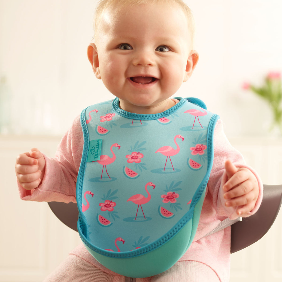 Baby wearing a Bibetta turquoise weaning bib with a flamingos pattern, made of neoprene material, with the pocket flipped out to the front, sitting in a highchair wearing a pink long sleeved t-shirt and leggings 