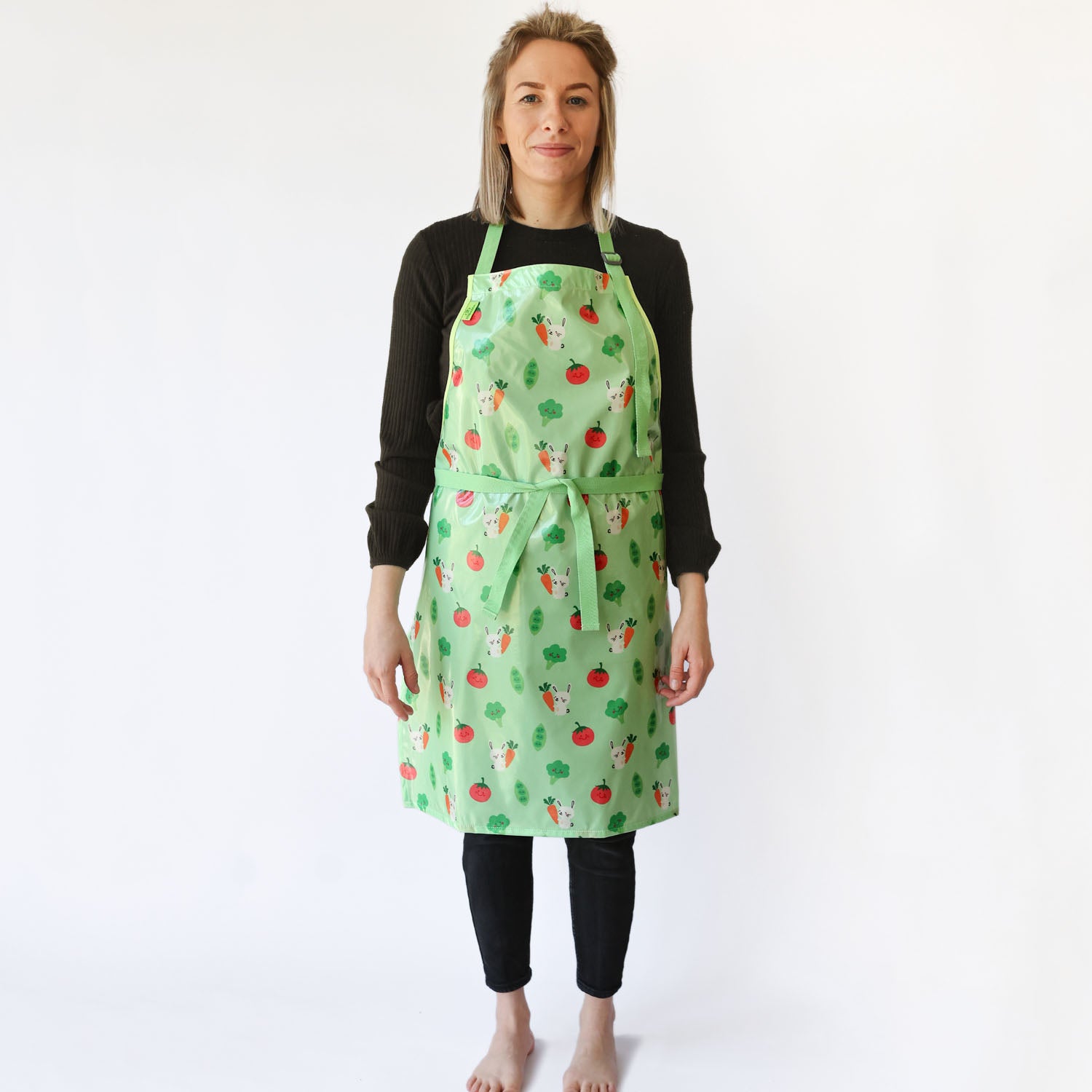 a smiling lady with blonde hair standing wearing a Bibetta Wipeezee adult apron in green bunny and veg pattern. She has fastened the waist straps at the front of the apron . She is wearing a black top and leggings and no shoes or socks.