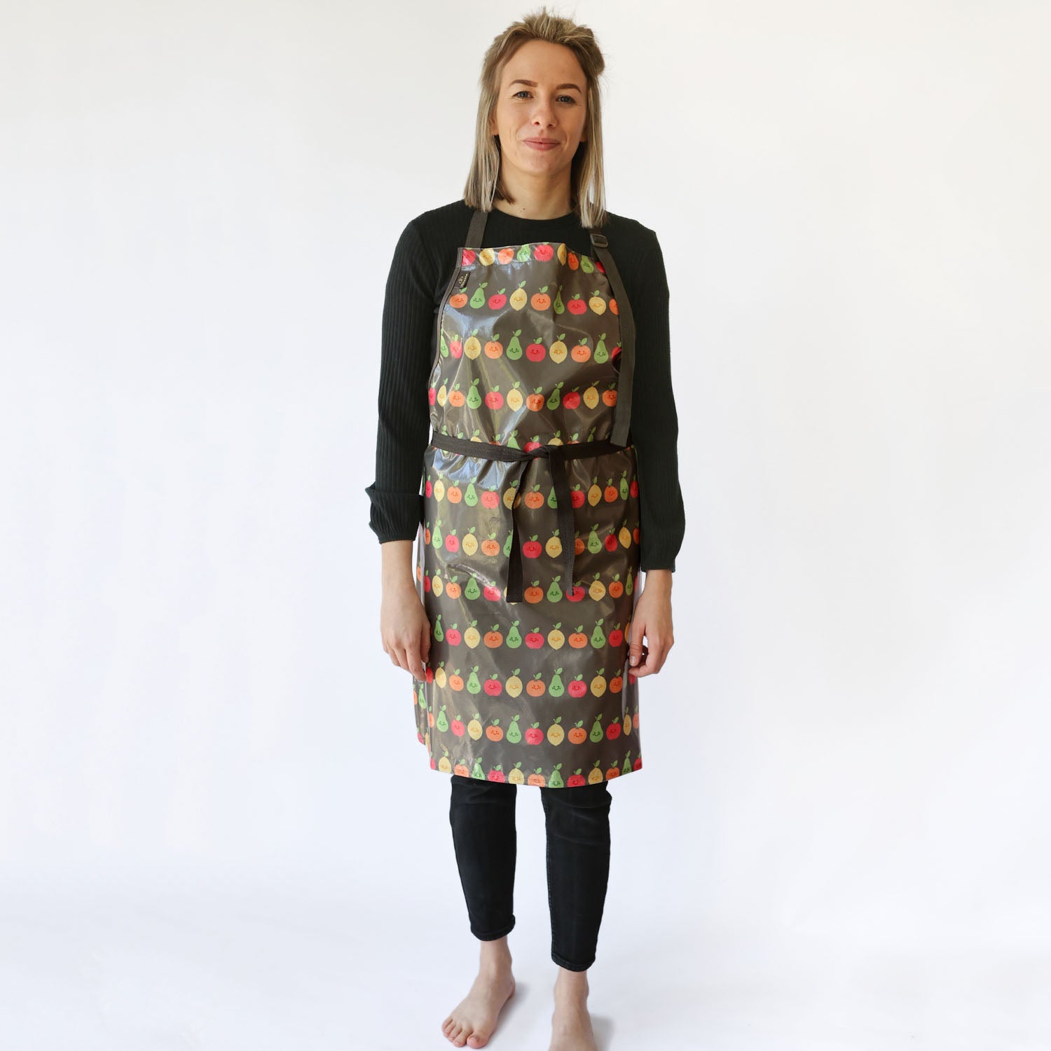 a smiling lady with blonde hair standing wearing a Bibetta Wipeezee adult apron in grey happy fruit pattern. She has fastened the waist straps at the front of the apron . She is wearing a black top and leggings and no shoes or socks.