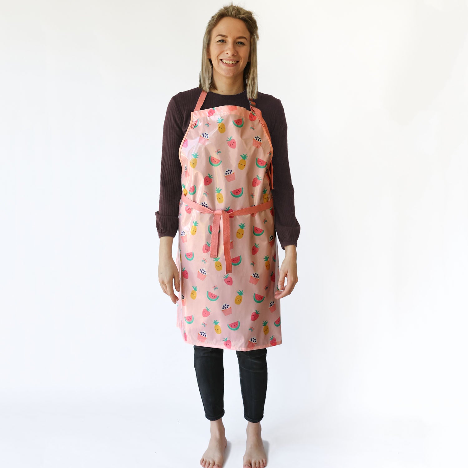 a smiling lady with blonde hair standing wearing a Bibetta Wipeezee adult apron in pink yummy treats pattern. She has fastened the waist straps at the front of the apron . She is wearing a black top and leggings and no shoes or socks.